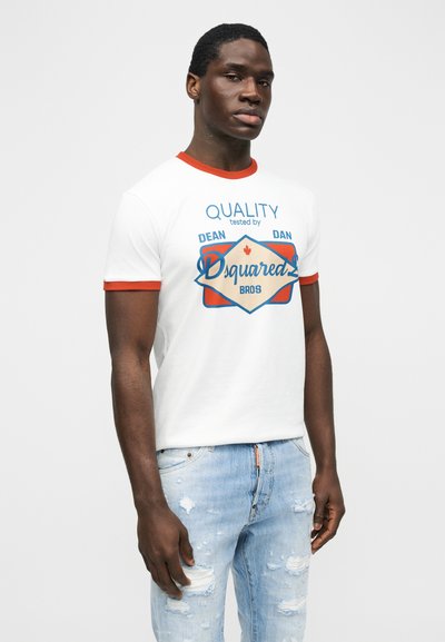Homme noir portant un t-shirt graphique blanc avec bordure rouge, logo bleu et beige, assorti à un jean bleu clair déchiré, debout devant un fond uni.
