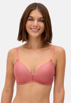 Mujer con cabello castaño hasta los hombros sonriendo, vistiendo un sujetador rosa texturizado con tirantes finos sobre un fondo liso.