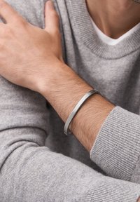 Bracelet métallique argenté sur un poignet avec des poils d'avant-bras châtain clair, porté avec un pull gris avec des poignets et un col côtelés.