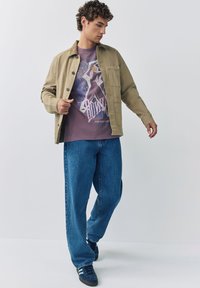 Man in beige jas over een paarse shirt met print, ruime blauwe spijkerbroek en blauwe sneakers, poserend tegen een effen lichtgrijze achtergrond.