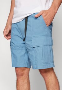 Man med ljusblå cargoshorts med flera fickor och svart dragsko, hand i höger ficka, vit skjorta delvis synlig.