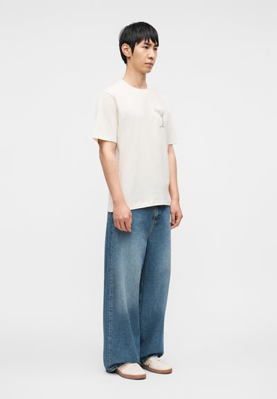 Κοντομάνικο κρεμ μπλουζάκι με γραφικό τύπωμα και τζιν παντελόνι με φαρδιά μπάρα σε μπλε χρώμα. Άσπρα αθλητικά παπούτσια με καστανές σόλες ολοκληρώνουν το ντύσιμο.