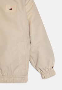 Beige Jacke mit einer weichen, glatten Textur. Verfügt über elastische Bündchen und eine Tasche, mit einem Tommy Hilfiger Logo-Patch im oberen Bereich.
