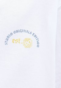 Balts džemperis ar zilu un dzeltenu izšūtu tekstu: "STUDIO ORIGINALS EDITION" un "est. 90" priekšpusē. Mīksts kokvilnas materiāls.