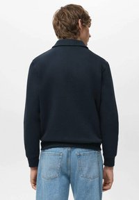 Granatowa, długi rękaw, polo z kołnierzykiem, ściągaczami na rękawach i dole; noszone z jasnoniebieskimi dżinsami, widziane od tyłu.