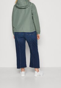 Light green, hooded jacket with elastic cuffs worn over dark blue wide-leg jeans and white sneakers. Smooth texture, casual design.