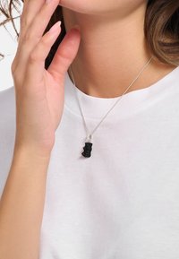 Schwarzer Anhänger in abstrakter Form an einer Silberkette, getragen über einem weißen Hemd. Die Halskette verfügt über eine strukturierte Oberfläche, die für visuelles Interesse sorgt.