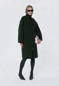 Kvinde iført en mørkegrøn frakke, sorte strømpebukser, sorte hæle, sorte solbriller, prikket tørklæde, holder en lyserød håndtaske.