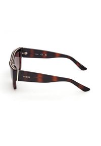 Lunettes de soleil en écaille avec des branches larges, des verres foncés, un détail chaîne dorée sur le dessus et le logo "GUESS" sur la branche.