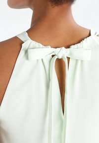 Světl zelený top bez rukávů s otvorem ve tvaru klíče na zádech, nastavitelné šnurování a nařasený výstřih z jemné, hladké látky.