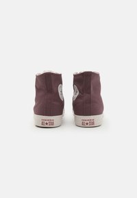 Tenisi înalți din pânză violetă cu tălpi de cauciuc alb și logo-ul roșu "CONVERSE ALL STAR". Au un design clasic cu glezna rotundă.