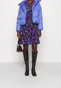 Blaue Pufferjacke, schwarzes und lila gemustertes Kleid, knielange schwarze Stiefel und eine kleine schwarze Handtasche. Fokus auf lebendige Farben und Texturen.