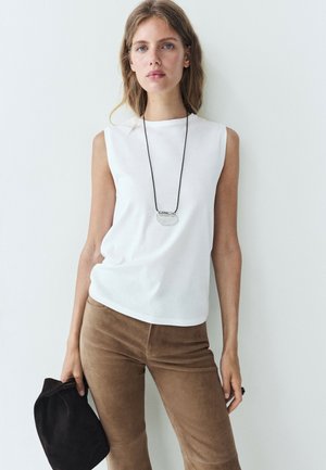 Jeune femme aux longs cheveux portant un top blanc sans manches, un pantalon marron, un collier pendentif, et tenant un chapeau noir contre un mur blanc.