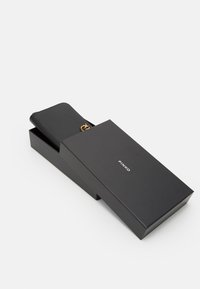 Portafoglio in pelle nera con un accento in hardware dorato, parzialmente visibile in una scatola nera con il nome del marchio "PINKO" sul fronte.