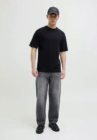 Joven con una gorra negra, camiseta negra de manga corta, jeans gris deslavado y zapatos negros, de pie contra un fondo liso claro.