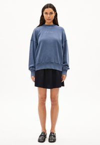 Sudadera de algodón azul con un corte relajado, puños acanalados y un pequeño logo en el pecho, combinada con una falda negra y mocasines negros.