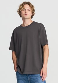 T-shirt in cotone grigio scuro con una vestibilità ampia, scollo a giro e maniche corte, abbinato a jeans azzurri chiari. Design minimalista, texture liscia.