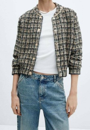 Femme portant une veste à carreaux texturée noire, blanche et beige boutonnée sur un débardeur blanc côtelé avec un jean bleu clair taille haute.