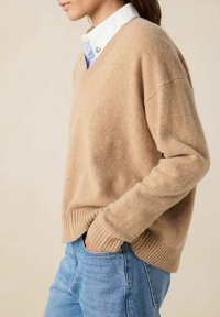 Pull en V beige en tricot doux avec un ourlet côtelé. Porté par-dessus une chemise blanche avec des accents bleus. La main du modèle dans la poche d'un jeans bleu clair.