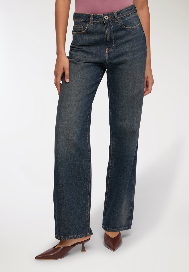 Jeans a gamba larga blu in un lavaggio scuro, con una vita alta, cinque tasche e dettagli di cucitura arancione, abbinati a mule con tacco marroni.