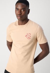 Beige Baumwoll-T-Shirt mit kurzen Ärmeln, Rundhalsausschnitt und einem pinken Grafiklogo auf der linken Brust. Glatte Textur, normale Passform.