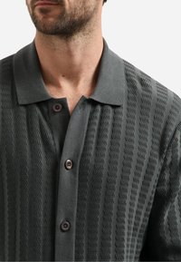 Donkergrijze gebreide polo met een geweven ontwerp met zigzagpatronen. Het heeft een kraag en bruine knopen.