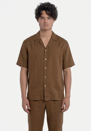 Uomo in piedi, indossa una camicia marrone a maniche corte con bottoni e pantaloni marroni abbinati su uno sfondo bianco semplice.