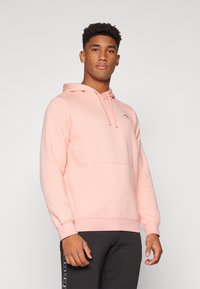 Haut à capuche rose clair avec cordon de serrage, poche kangourou et poignets côtelés. Porté sur un pantalon gris foncé, arborant un petit logo sur la poitrine.
