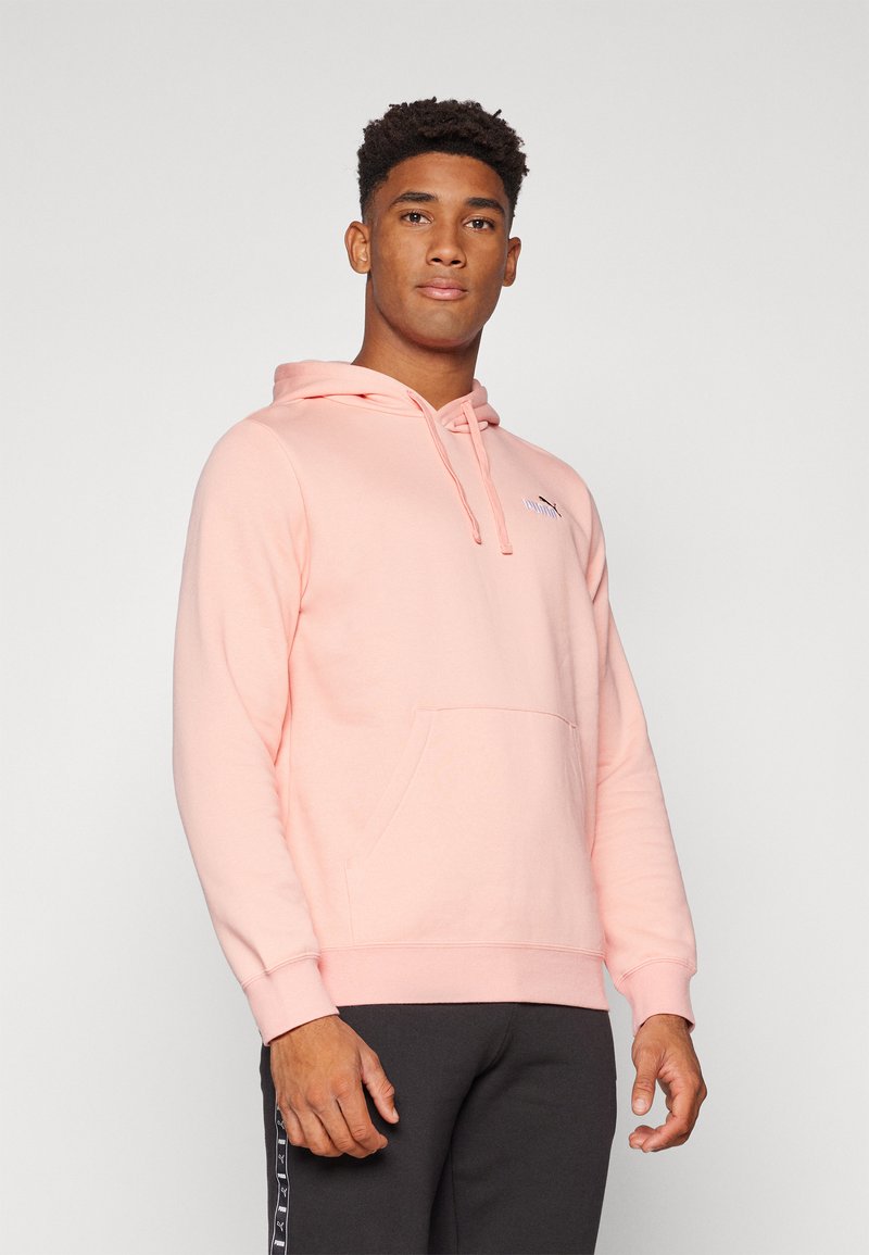 Haut à capuche rose clair avec cordon de serrage, poche kangourou et poignets côtelés. Porté sur un pantalon gris foncé, arborant un petit logo sur la poitrine.