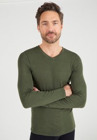 Olijfgroen langarm V-hals shirt van zachte stof, met een getailleerd ontwerp en een geribbelde manchet, gedragen door een man met gekruiste armen.