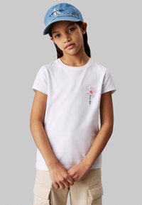 Camiseta de algodón blanca con mangas cortas, que presenta un diseño bordado floral en rosa y rojo en el lado izquierdo. Pantalones cargo beige y gorra de mezclilla.