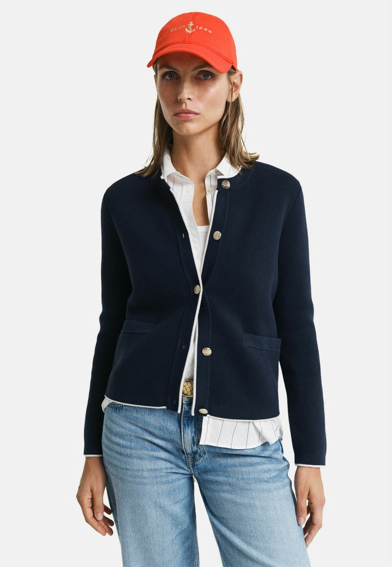 Marineblaue Strickjacke mit goldenen Knöpfen, zwei vorderen Taschen, über einem weißen Hemd mit Kragen getragen, kombiniert mit hellblauen Jeans und einer orangefarbenen Mütze.