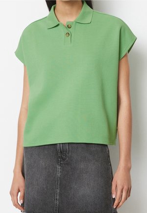 Frau trägt ein kurzärmeliges, hellgrünes Poloshirt kombiniert mit einem hoch taillierten schwarzen Jeansrock vor weißem Hintergrund.