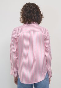 Person mit lockigem braunem Haar, von hinten sichtbar, trägt ein pinkes und weißes, senkrecht gestreiftes Langarmshirt und hellblaue Jeans.