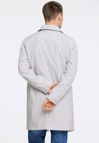 Trench coat grigio chiaro con colletto classico, maniche lunghe e polsini con bottone, indossato su jeans blu; materiale liscio e resistente all'acqua.