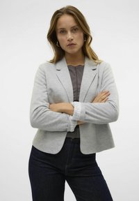 Blazer gris de tela suave con un diseño ajustado, que presenta solapas con muescas y puños enrollados sobre una camiseta más oscura y jeans oscuros.