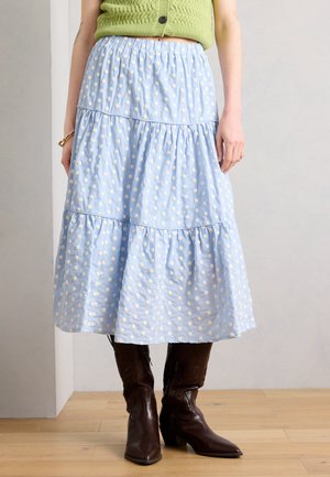 Žena má na sebe svetlomodrú viacvrstvovú midi sukňu s bielymi bodkami, zelený pletený top a tmavohnedé kožené čižmy na opätku na drevenej podlahe.