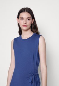 Vestido sem mangas azul com decote redondo, textura franzida de um lado e detalhe de laço, feito de material macio e elástico.