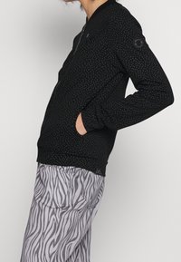 En person som bär en svart zip-up jacka med små vita prickar och grå byxor med ett zebramönster, händerna i jackfickorna.