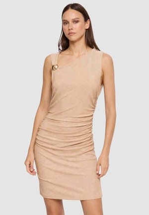 Donna che indossa un mini abito beige senza maniche, arricciato, con una scollatura asimmetrica chiusa da un grande bottone dorato su una spalla.