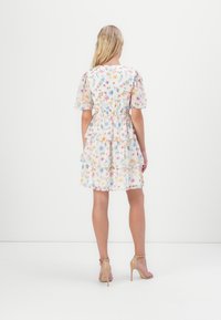 Robe à imprimé floral avec des manches bouffantes, taille froncée, jupe à volants et tissu transparent dans des couleurs pastel. Portée avec des sandales à talons nus.