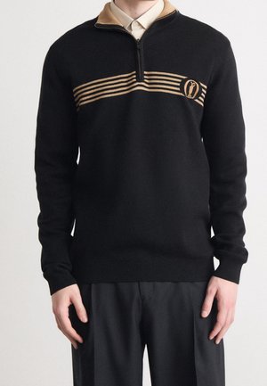 Mann trägt schwarzen Pullover mit hellbraunem Kragen, Reißverschluss und waagrechten Streifen über der Brust, kombiniert mit schwarzer Hose.