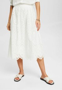 Nainen, jolla on päällä valkoinen eyelet-midi-hame, valkoinen pusero ja valkoiset liukuvapohjaiset sandaalit, joissa on ristiin menevät hihnat, yksinkertaisella taustalla.