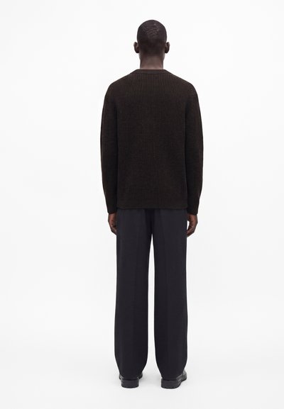Homme aux cheveux courts portant un pull en maille côtelée marron foncé, un pantalon large noir et des chaussures noires, debout dos à un fond blanc.