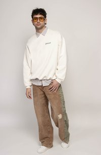 Kremgenser med teksten "Good Luck", lagd over en stripete skjorte. Brune vide bukser med slitte detaljer og grønn sideststripe. Hvite sneakers.