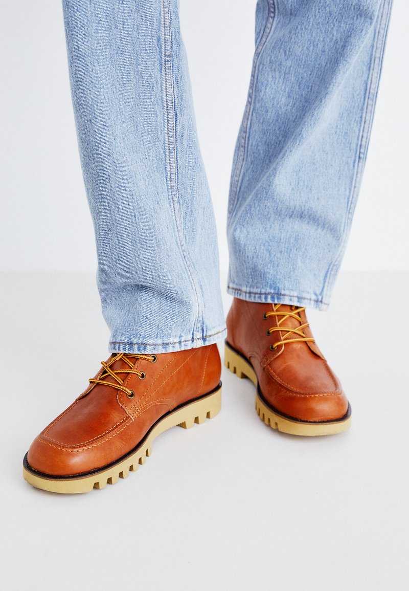 Botas de cuero marrón con cordones amarillos y una suela beige resistente. Llevadas con jeans de mezclilla de color azul claro, mostrando detalles de costura en las botas.