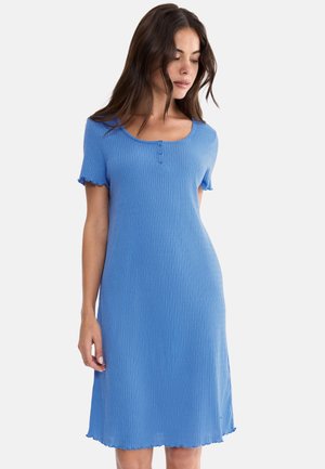 Kobieta ubrana w niebieską, prążkowaną sukienkę z krótkim rękawem, z trzema guzikami na piersi i falbanami na krawędziach, stojąca na białym tle.