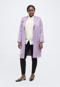 Γυναίκα στέκεται μπροστά σε λευκό φόντο φορώντας λιλά παλτό, κρεμ πουκάμισο με βολάν, μαύρο παντελόνι και μαύρα παπούτσια με χαμηλό τακούνι.