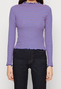 Paski długi rękaw w kolorach różowym i niebieskim z prążkowaną fakturą. Posiada falbaniaste wykończenie dekoltu i dołu, zestawiony z ciemnymi dżinsami.