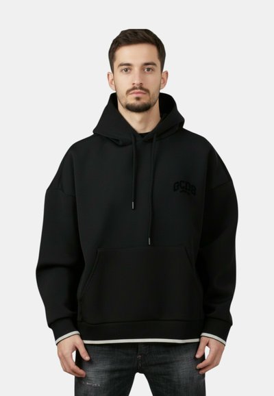 Jeune homme aux cheveux courts et foncés portant un sweat à capuche noir avec une poche avant et un logo discret "GCDS", associé à un jean foncé.